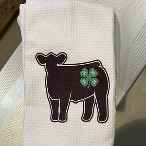 May include: White kitchen towel with a black silhouette of a cow with a green four-leaf clover with the letters "H" on each leaf. The clover is on the cow's side.