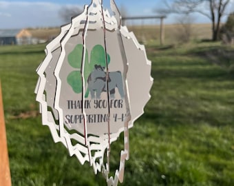 Può includere: Un ornamento argentato a strati con un bordo smerlato è tenuto contro un cielo blu. L'ornamento presenta un quadrifoglio verde, una grafica di mucca e il testo "THANK YOU FOR SUPPORTING 4-H".