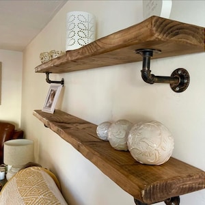 May include: Two rustic wooden shelves with black metal pipe brackets. The shelves are mounted on a white wall and decorated with decorative objects, including a picture frame, a candle holder, and decorative balls.