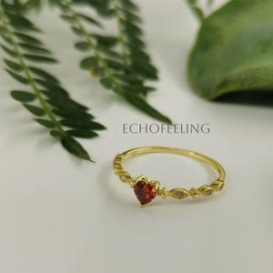 May include: A gold ring with a heart-shaped red gemstone at the center. The ring is surrounded by small, clear gemstones and decorative elements. The word "ECHOFEELING" is visible in the background.