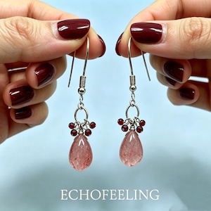 May include: A pair of dangle earrings with silver hooks. Each earring features a cluster of small red beads above a teardrop-shaped, pinkish-red gemstone. The earrings are held up by a person's hands. The word "ECHOFEELING" is at the bottom.