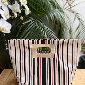 Puede incluir: Un bolso tote rectangular de tela con un patrón de rayas verticales en negro, rojo y blanco roto. El bolso incluye una abertura rectangular beige tipo cuero para el asa, integrada directamente en el borde superior del cuerpo textil.