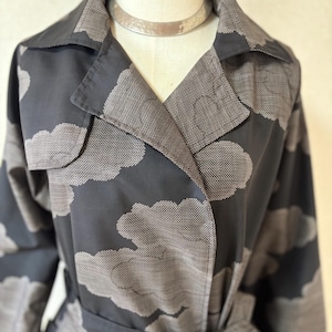 Op de afbeelding: Een zwarte trenchcoat met een herhalend patroon van grijze wolken met een gestippeld textuurontwerp. De jas heeft een brede reverskraag, lange mouwen en een bijpassende stoffen ceintuur die in de taille is gestrikt, getoond op een paspop.