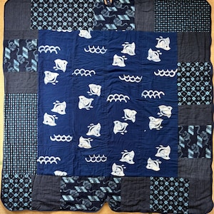 May include: A square patchwork quilt featuring a central dark blue panel with a repeating white print of stylized fish and wave patterns. The border consists of various dark blue and light blue fabric squares with geometric and floral motifs.