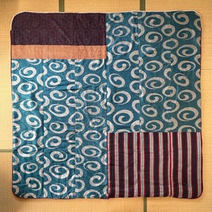 May include: A rectangular quilted blanket featuring a patchwork design. The fabric includes sections with a teal and silver spiral pattern, a dark purple floral print, a thin orange and white checkered band, and a dark striped section with red lines.