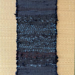 Puede incluir: Una alfombra tejida rectangular que presenta una paleta de colores azul oscuro y negro con sutiles toques de rojo y azul claro. El tejido tiene una textura tipo trapo con bordes deshilachados y flecos de color blanco roto en los extremos.