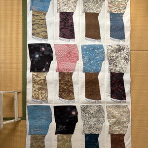 May include: A fabric quilt featuring fifteen stylized kimono designs. Each kimono has a unique pattern and color scheme, including floral and geometric prints in shades of blue, pink, brown, and black. The quilt is displayed on a tatami mat.