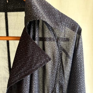 以下が含まれることがあります： 小さな菱形と十字が繰り返される白い幾何学模様が施された、ダークグレーの半透明のボタンダウンシャツ。この衣類は尖った襟が特徴で、木製のハンガーに掛けられており、軽量で質感のある生地の様子が確認できます。