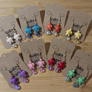 May include: A collection of colorful turtle earrings displayed on brown tags. The earrings feature small turtle charms in various colors, including white, blue, orange, yellow, pink, red, purple, and green, with dangling beads. The earrings are on gold-colored hooks.