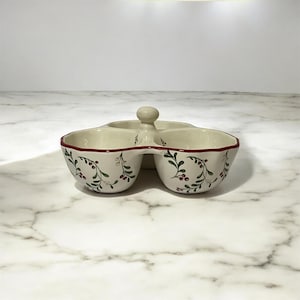 May include: A three-section ceramic serving dish with a central handle. The off-white exterior features a painted pattern of green leaves and red berries. The rim of each bowl is accented with a thin red line, and the interior is smooth and cream-colored.