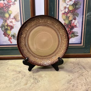 May include: A decorative ceramic plate with a brown rim and a tan center. The plate features a geometric pattern around the edge. It is displayed on a black stand, with framed artwork in the background.