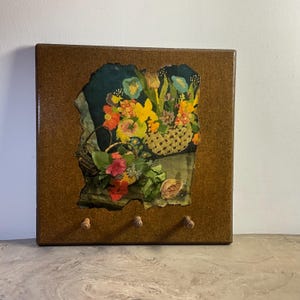 May include: A square brown wooden wall hook rack featuring a central inset panel with a colorful floral print. The print depicts a basket of flowers in shades of red, yellow, and green against a dark background. Three small wooden pegs are attached.