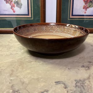 May include: A brown ceramic bowl with a decorative geometric pattern around the rim. The bowl has a wide, shallow shape and a glossy finish. The interior is a lighter shade of brown, and the bowl sits on a marble surface.