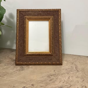 May include: A rectangular picture frame with a brown, textured border and a gold inner frame. The frame has an ornate, embossed design. The background is white, and the frame is sitting on a marble-like surface.
