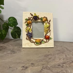 May include: Square cream-colored picture frame with a decorative border of sculpted fruit and vines. The frame features grapes, pears, and an apple. The frame is made of a textured material and has a rectangular opening for a photo.
