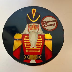 May include: Round tin with a navy blue background featuring a nutcracker soldier design. The soldier wears a red coat with gold buttons and a black hat. The tin reads "Shortbread Cookies" in a circular label.