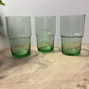 May include: Three clear, stackable drinking glasses with a light green tint. The glasses have a cylindrical top and a smaller, rounded base, creating a unique, modern design. The glasses are empty and sit on a light-colored surface.
