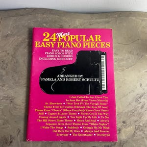 May include: A music book titled 24 More Popular Easy Piano Pieces with a bright pink cover. The cover features a black grand piano graphic, text stating it is arranged by Pamela and Robert Schultz, and a list of song titles in yellow text.