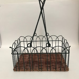 May include: A hanging basket with a dark green metal frame and a woven brown base. The frame has a scalloped edge design. The basket is suspended by two twisted metal chains.