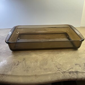 May include: A rectangular baking dish made of translucent brown glass. The dish features a smooth, open-top design with rounded corners and a wide, flat rim extending around the entire perimeter, providing a clear view of the interior base.