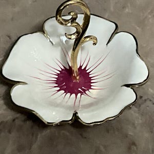 May include: A white, flower-shaped dish with a gold-colored handle. The dish has a gold trim and a deep magenta center with magenta lines radiating outwards. The handle is a decorative, curved design.