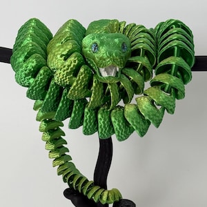 May include: A green snake figurine coiled around a black tree branch. The snake has a textured surface and blue eyes. The tree branch has a twisted, gnarled shape.