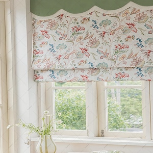 Puede incluir: Una cortina romana con estampado floral y borde festoneado está montada sobre una ventana. La cortina presenta un fondo crema con diseños florales rojos, azules y verdes. Una cenefa verde remata la cortina. Una pequeña mesa con un jarrón y un libro está frente a la ventana.
