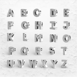 May include: Silver alphabet charms, each letter encrusted with small, clear crystals. The letters A through Z are displayed, showcasing a variety of fonts and designs. These charms are likely for bracelets or necklaces.