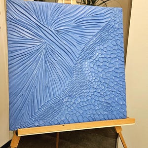 Può includere: Opera d'arte blu strutturata su cavalletto in legno. La tela quadrata presenta una combinazione di texture lineari e a motivi. L'opera è esposta in interni, con una pianta visibile sullo sfondo. Dimensioni approssimative: 76 cm x 76 cm.