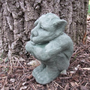 May include: A small, weathered, green gargoyle statue in a squatting position. The statue has large ears and is positioned in front of a tree trunk, surrounded by leaves and twigs, suggesting an outdoor garden setting.