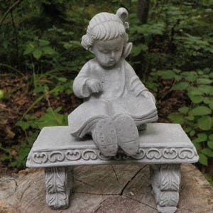 May include: A gray concrete statue of a child reading a book while seated on a bench. The statue has detailed features, including the child's clothing and the bench's decorative trim. The statue is outdoors, surrounded by greenery.
