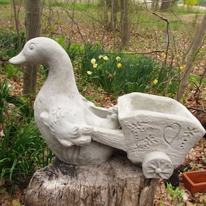 May include: A concrete duck planter with a cart attached, resting on a tree stump. The cart features floral and heart designs. The duck is light gray, and the scene is set outdoors with greenery.