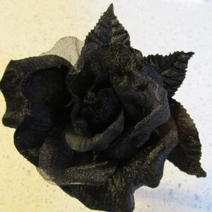 May include: A black fabric rose with detailed petals and leaves. The flower is a dark, solid color, and the petals have a textured appearance. The rose is a decorative item, possibly for crafts or embellishment.