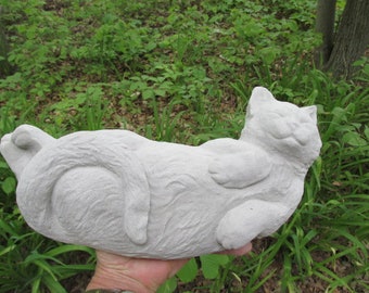May include: A light gray cat sculpture in a relaxed pose, lying on its back with paws up. The cat's fur is textured, and its eyes are closed, giving it a peaceful expression. The sculpture is held in a hand against a backdrop of green foliage.