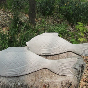 May include: Two light gray concrete fish sculptures with textured scales and fins. The fish are lying on a tree stump, with a natural outdoor background of green foliage and trees.