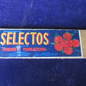May include: A rectangular wooden label with the words "SELECTOS BRAND CHERRY TOMATOES" in yellow and red lettering on a blue background. A cluster of red cherry tomatoes with green leaves is on the right side. The label reads "Grown in Mexico".