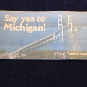 May include: A vintage Michigan transportation map with the text "Say yes to Michigan!" in gold lettering. The map features a bridge over water and the words "Official Transportation Map".