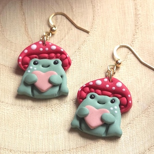 May include: Handmade earrings with a charming design. Each earring depicts a light green frog holding a pink heart, under a red mushroom cap with white spots. The earrings have gold-coloured ear wires.