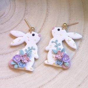 Op de afbeelding: Een paar witte konijnenoor-oorbellen met pastelroze, blauwe en groene bloemmotieven. De oorbellen zijn gemaakt van polymer clay en hebben gouden haakjes.