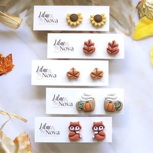 May include: A selection of autumn-themed stud earrings. Designs include sunflowers, leaves, pumpkin spice lattes, and owls. Each pair is displayed on a white card with the text "Lilac & Nova". The earrings are in shades of brown, orange, and yellow.