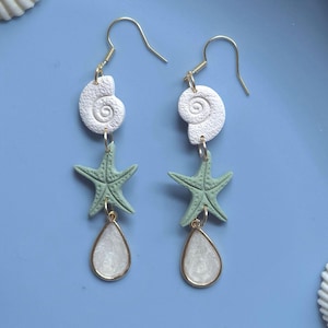 Peut inclure: Boucles d'oreilles pendantes sur le thème de la plage. Chaque boucle d'oreille est ornée d'un coquillage blanc, d'une étoile de mer verte et d'un pendentif en forme de goutte d'eau avec un cadre doré. Les boucles sont suspendues à des crochets dorés.