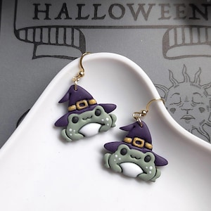May include: A pair of dangle earrings featuring sculpted green frogs wearing purple witch hats with gold buckles. The frogs have white bellies and dark eyes, suspended from gold-toned metal hooks. The design is detailed and colourful.