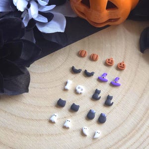 May include: A collection of Halloween-themed miniature charms in various shapes, including pumpkins, bats, witch hats, cats, ghosts, and cauldrons. The charms are in orange, black, white, and purple colors, arranged on a wooden surface.
