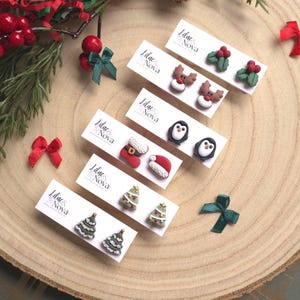 Peut inclure: Une collection de boucles d'oreilles clous sur le thème de Noël, présentées sur des cartes blanches. Les motifs incluent des rennes, des pingouins, des sapins, du houx, des bonnets de Père Noël et des bas de Noël. Décorations festives.