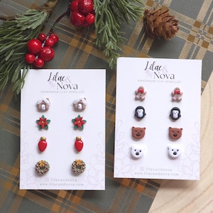 May include: Two cards of handmade clay jewellery from Lilac & Nova. One card features earrings shaped like houses, poinsettias, Christmas lights, and wreaths. The other card has gingerbread men, penguins, bears, and polar bears. The background is a plaid pattern.