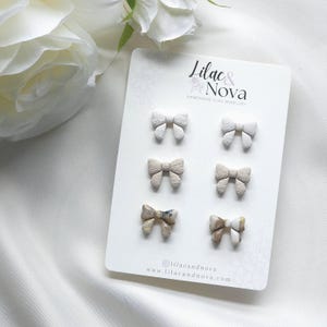 May include: A set of six handmade clay bow earrings in various shades of beige and white. The earrings are displayed on a white card with the text "Lilac & Nova Handmade Clay Jewellery" printed on it.