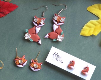 May include: Handmade fox earrings and studs. The earrings feature a fox design with orange, white, and brown colors, adorned with white flower accents. The studs and earrings are displayed on a green surface with fall-themed decorations.
