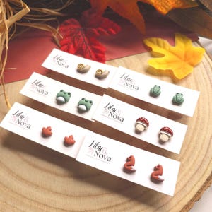 May include: Handmade clay earrings in various designs: snails, frogs, mushrooms, foxes, and leaves. Each pair is displayed on a white card with the "Lilac & Nova" logo. The earrings are set against a wooden background with autumn leaves.