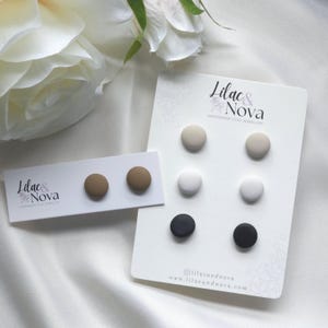 May include: Handmade clay stud earrings in shades of brown, beige, white, and black, displayed on white cards. The cards feature the "Lilac & Nova" logo and website. The earrings are a simple, minimalist design.
