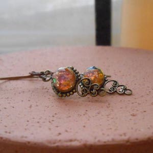 May include: An antique-style hair barrette with two iridescent orange-yellow cabochons set in ornate bronze-toned metal. The barrette is resting on a round, textured, pink surface. The design features filigree details.
